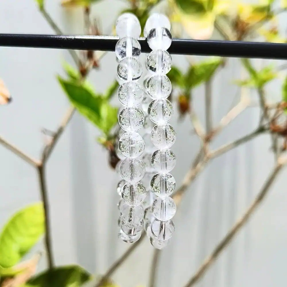 CRYSTCOP clear quartz bracelet with round transparent beads hanging on a branch with blurred natural background